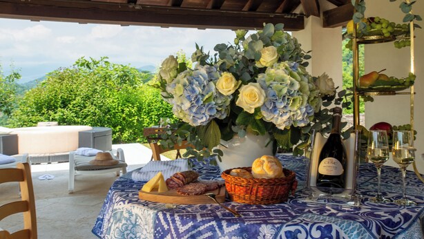 An outdoor dining area with a table set for two, featuring a bottle of wine, glasses, bread, and a basket of pastries. - Tenuta Castagnera (Tarzo)