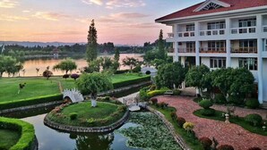 A multi-story hotel with a red roof, surrounded by a well-maintained garden with a circular water feature, a bridge, and a lake view.