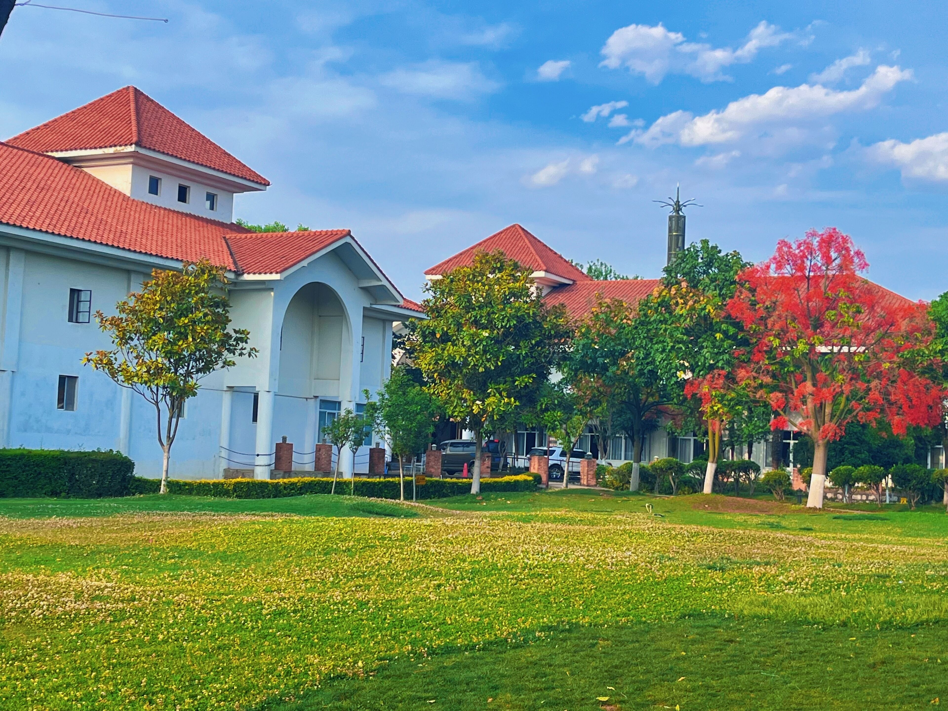 A large building with a red roof and white walls, surrounded by green grass and trees.