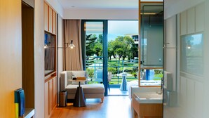 A modern hotel room with a balcony, a sofa, a small table, a trash bin, and a bathroom with a sink and mirror.
