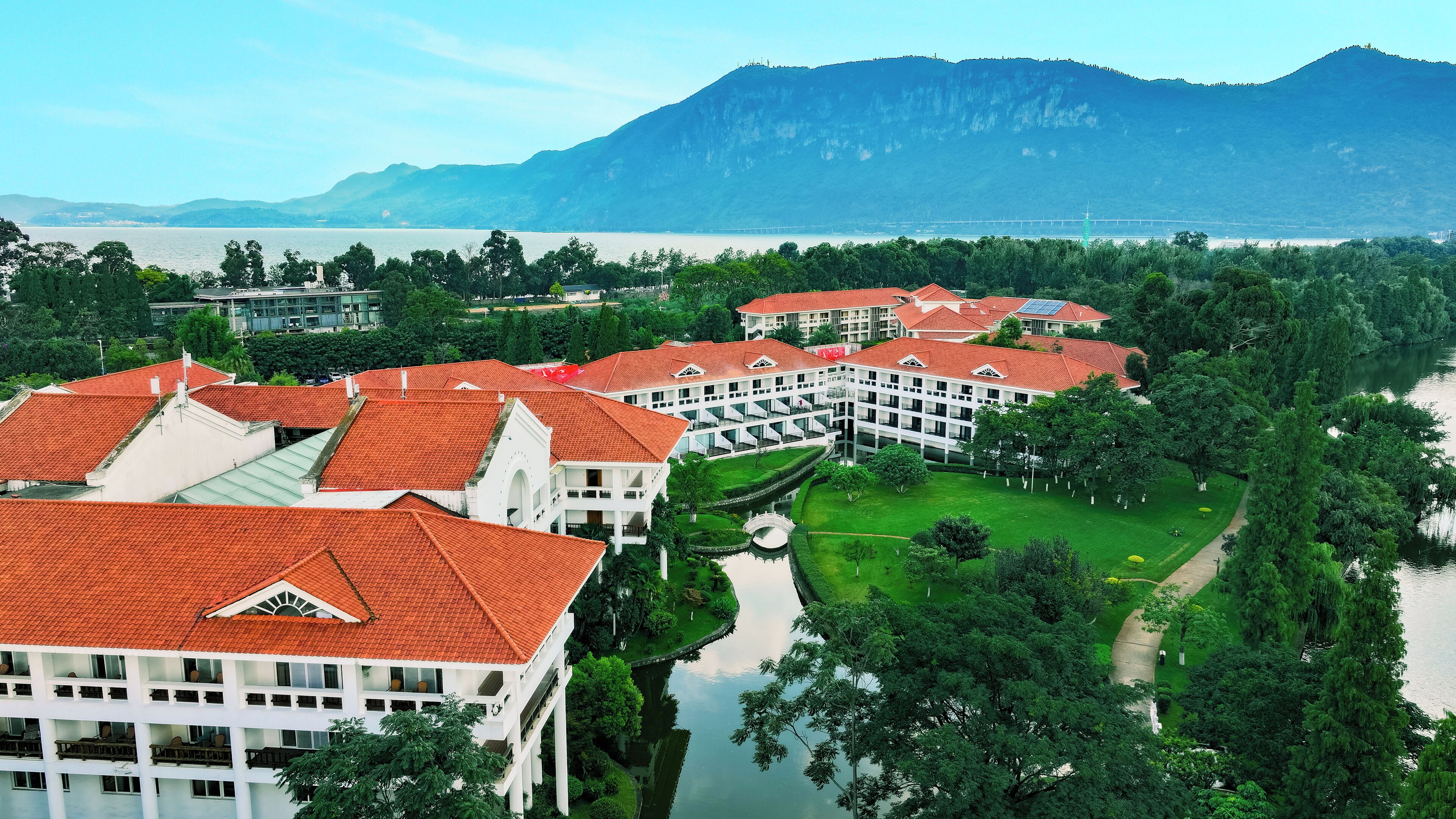 A large hotel with red-tiled roofs and multiple floors, surrounded by lush greenery and a body of water.