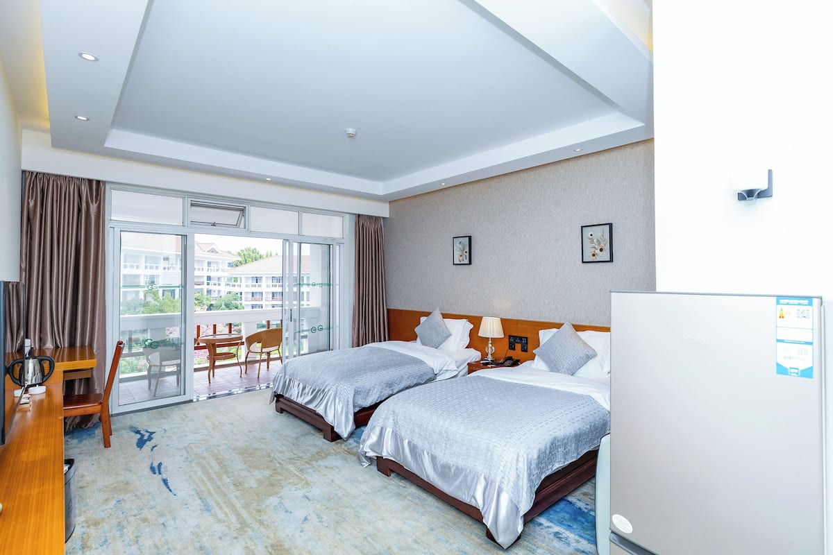 Ein Hotelzimmer mit zwei Betten, einem Schreibtisch, einem Stuhl, einem Kühlschrank und einem Balkon mit Blick auf Gebäude.