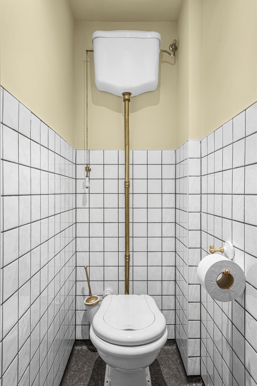 A small, tiled bathroom with a white toilet, a gold flush handle, and a white toilet paper holder.