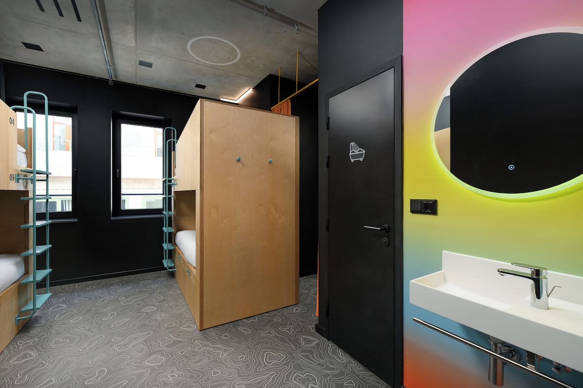 A modern hotel room with bunk beds, a wooden wardrobe, a round mirror with a neon light, and a white sink.