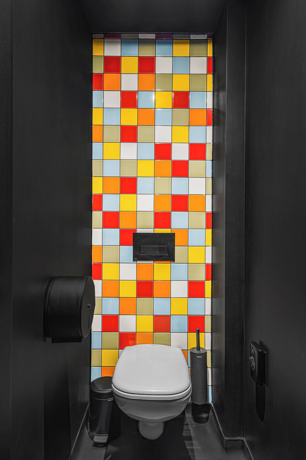 A small, modern bathroom with a colorful mosaic tile backsplash, a toilet, a trash can, and a black toilet paper holder.