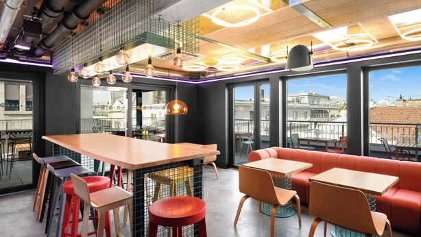 A modern interior with a bar area, seating, and large windows offering city views. - JO&JOE Budapest (Budapest)