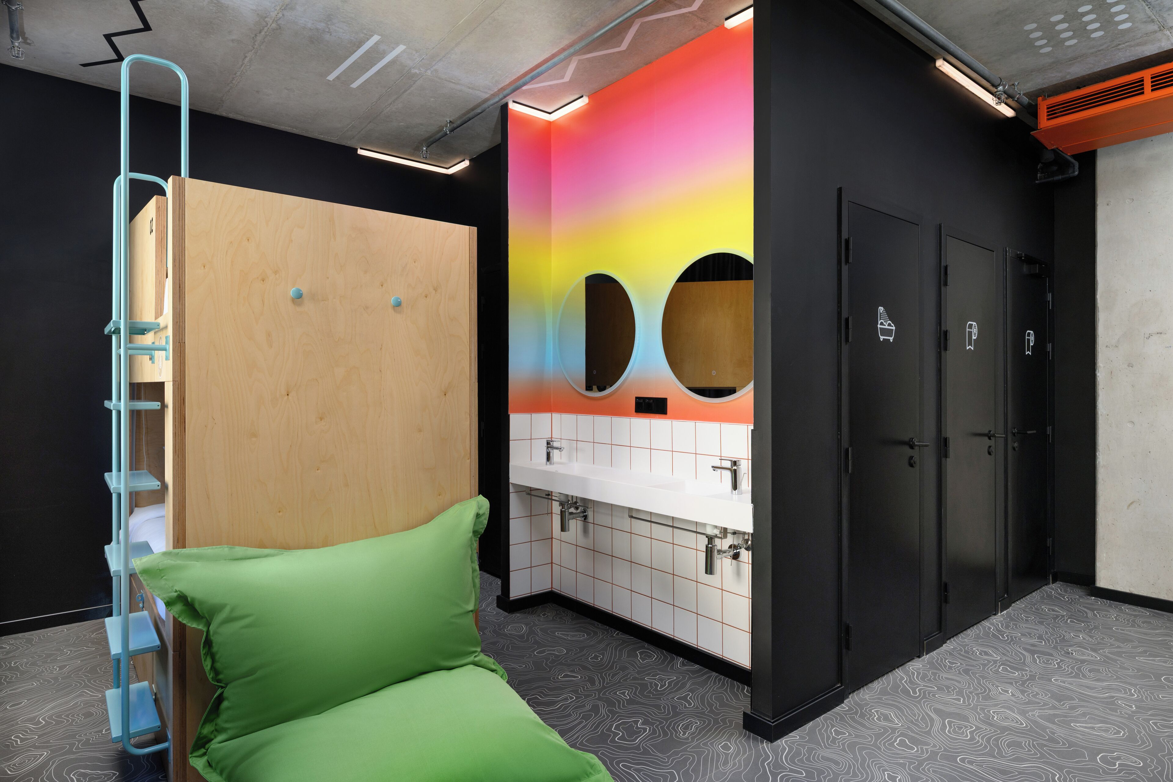 Un baño moderno con separador de madera, un banco acolchado verde, una pared colorida con orificios circulares y una zona de lavabo blanca.
