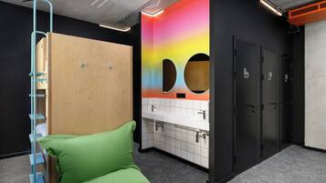 Un baño moderno con separador de madera, un banco acolchado verde, una pared colorida con orificios circulares y una zona de lavabo blanca.