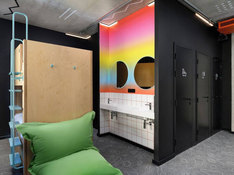 Un baño moderno con separador de madera, un banco acolchado verde, una pared colorida con orificios circulares y una zona de lavabo blanca.