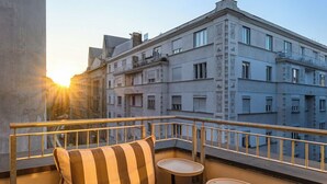 A balcony with striped cushions and small tables, overlooking a cityscape with buildings and a sunset. - JO&JOE Budapest (Budapest)