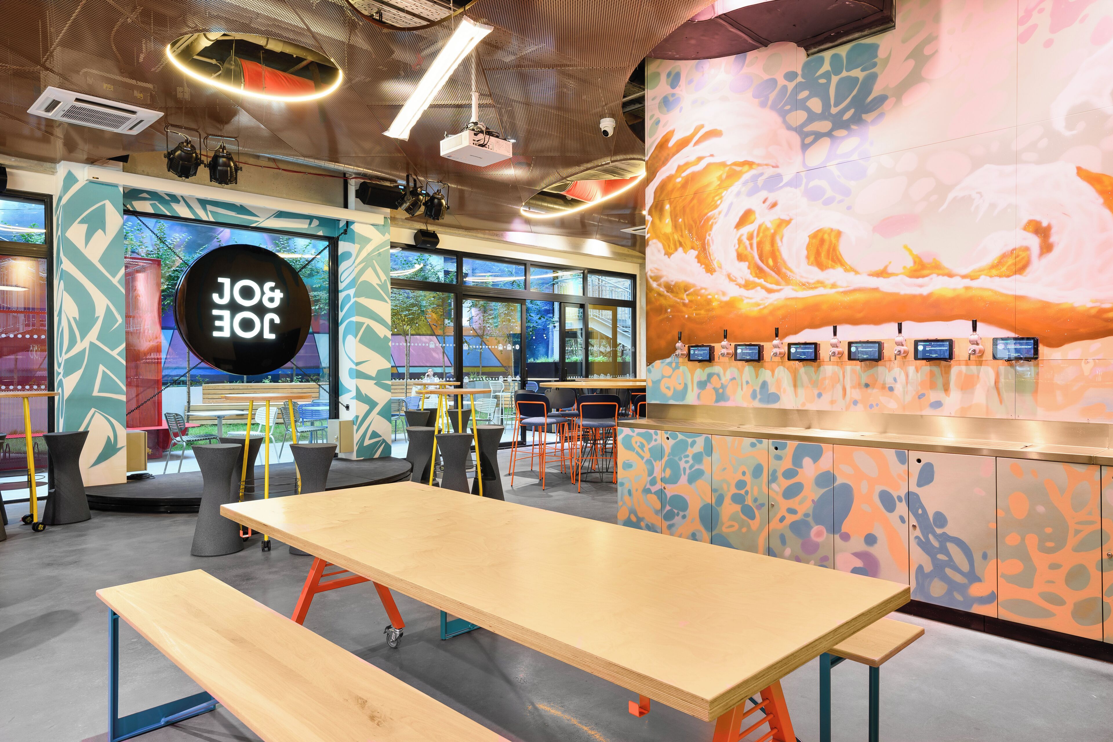 A modern cafe with a large wooden table, colorful abstract wall art, and a digital menu display.