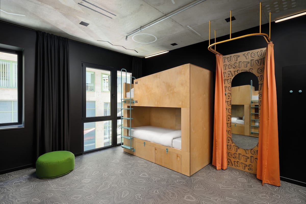 A bunk bed with a ladder, a green ottoman, and a window with curtains.