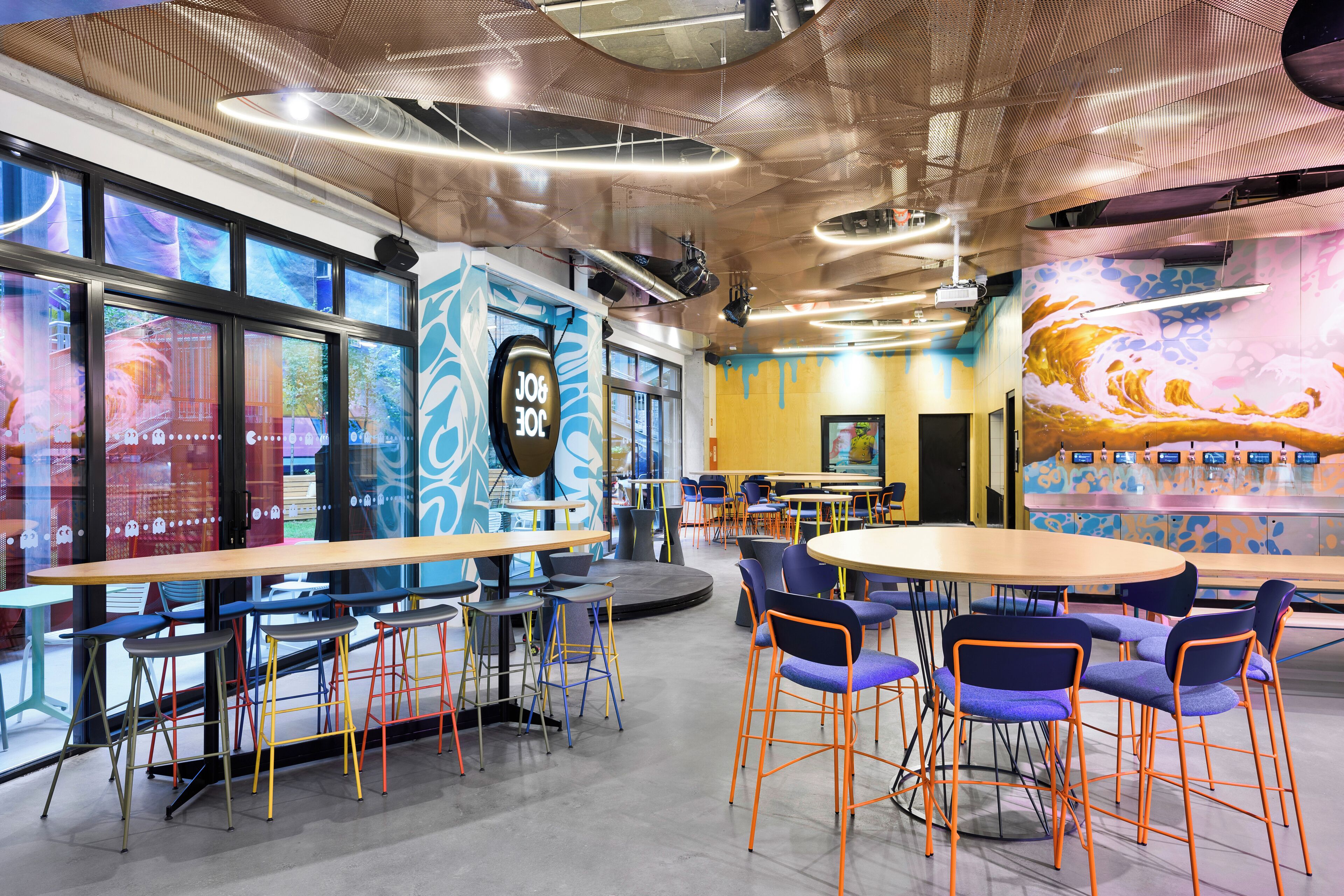 A modern indoor space with tables and chairs, a stage, and a colorful mural.