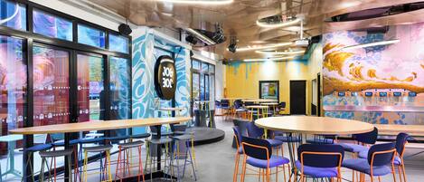 Un espacio interior moderno con mesas, sillas, un escenario y un mural colorido.
