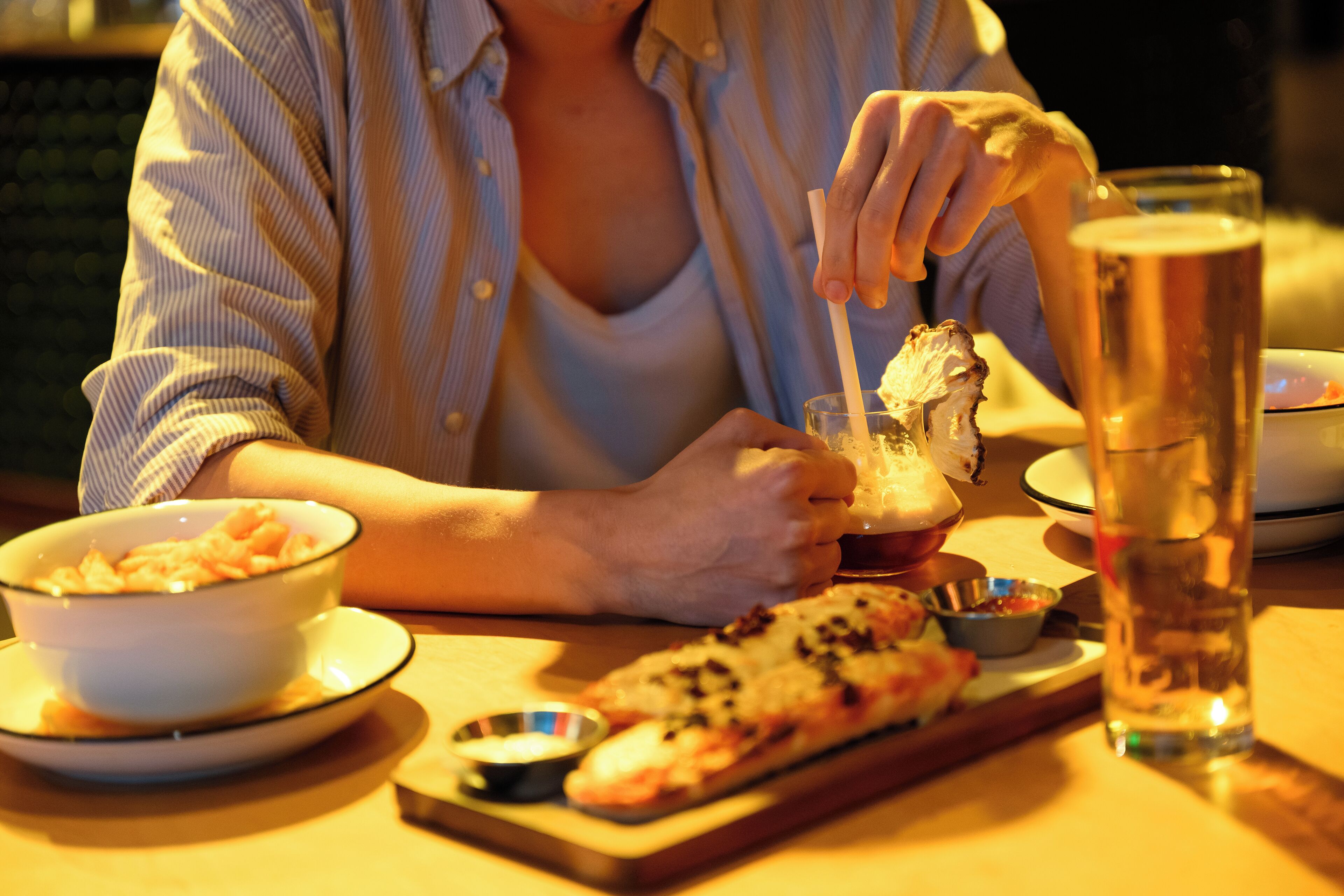 A person sitting at a table with a meal consisting of a bowl of pasta, a slice of pizza, and a glass of beer.