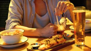 A person sitting at a table with a meal consisting of a bowl of pasta, a slice of pizza, and a glass of beer.