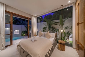 A modern bedroom with a large bed, a small wooden stool, and a view of a swimming pool. - Villa Lilu  (Canggu)