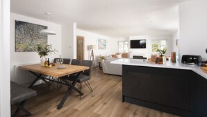 A modern kitchen with a central island, a dining area with a wooden table and chairs, and a living space with a sofa and a TV. - Tre Lowen- Enclosed Garden - Free Parking (Perranporth)