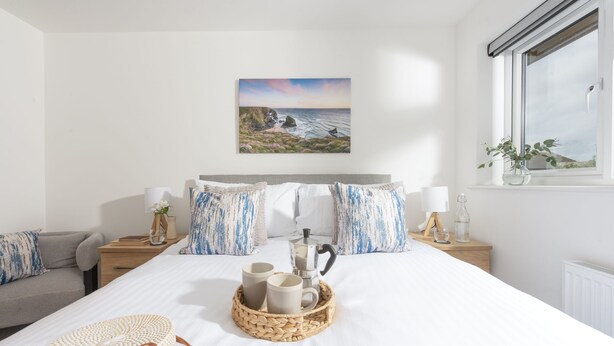 A neatly arranged bedroom with a bed, bedside tables, a window with a view, and a painting on the wall. - Tre Lowen- Enclosed Garden - Free Parking (Perranporth)