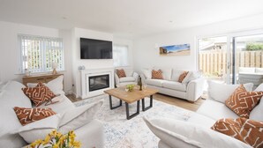 A spacious living room with a fireplace, white sofas, a coffee table, and a TV mounted on the wall. - Tre Lowen- Enclosed Garden - Free Parking (Perranporth)