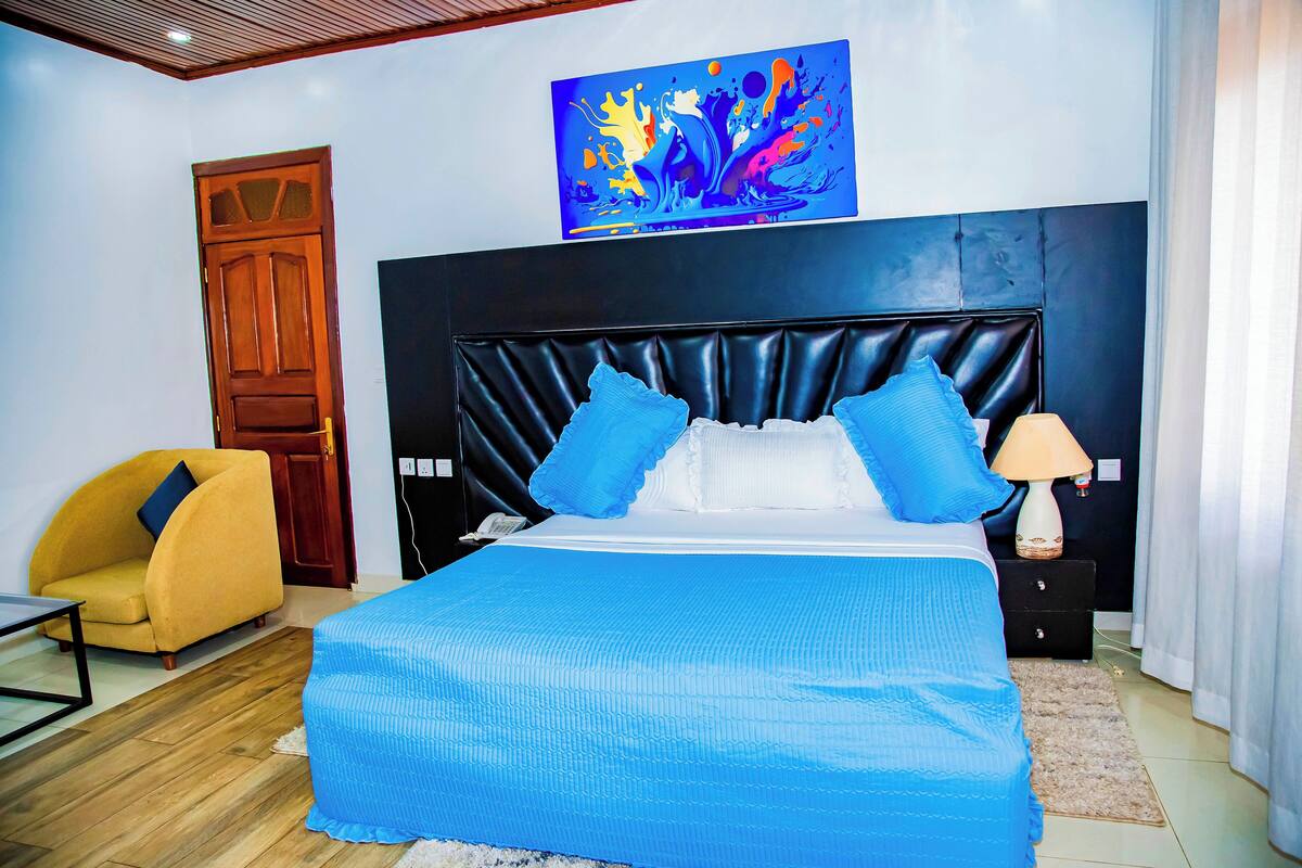 A bedroom with a large bed, a wooden door, a yellow armchair, and a painting on the wall.
