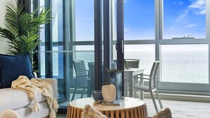 Un salon moderne avec un canapé, une table basse ronde en bois et une vue sur l’océan grâce à de grandes fenêtres.