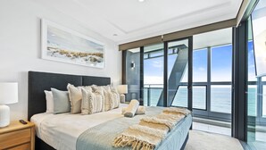 Une chambre moderne avec un grand lit, une table de chevet, une lampe et une vue sur l’océan grâce à de grandes fenêtres.