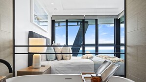 A modern hotel room with a large window, a bed with white linens, a bedside table with a lamp, and a bathtub with a tray.