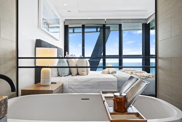 A modern hotel room with a large window, a bed with white linens, a bedside table with a lamp, and a bathtub with a tray.