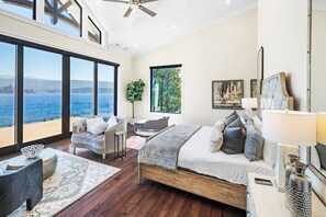 Ein Schlafzimmer mit großem Fenster und Blick auf ein Gewässer, einem Bett mit vielen Kissen, einem Nachttisch und einem Sessel.