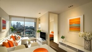 Un salon moderne avec vue sur la ville, un coin repas avec une table blanche et un téléviseur posé sur un meuble.