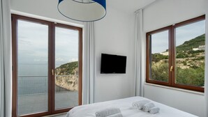 Ein Schlafzimmer mit Bett, Fernseher und großen Fenstern mit Blick auf Meer und Hügel.