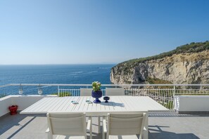 Une terrasse avec une table et des chaises blanches, donnant sur une falaise côtière et la mer.