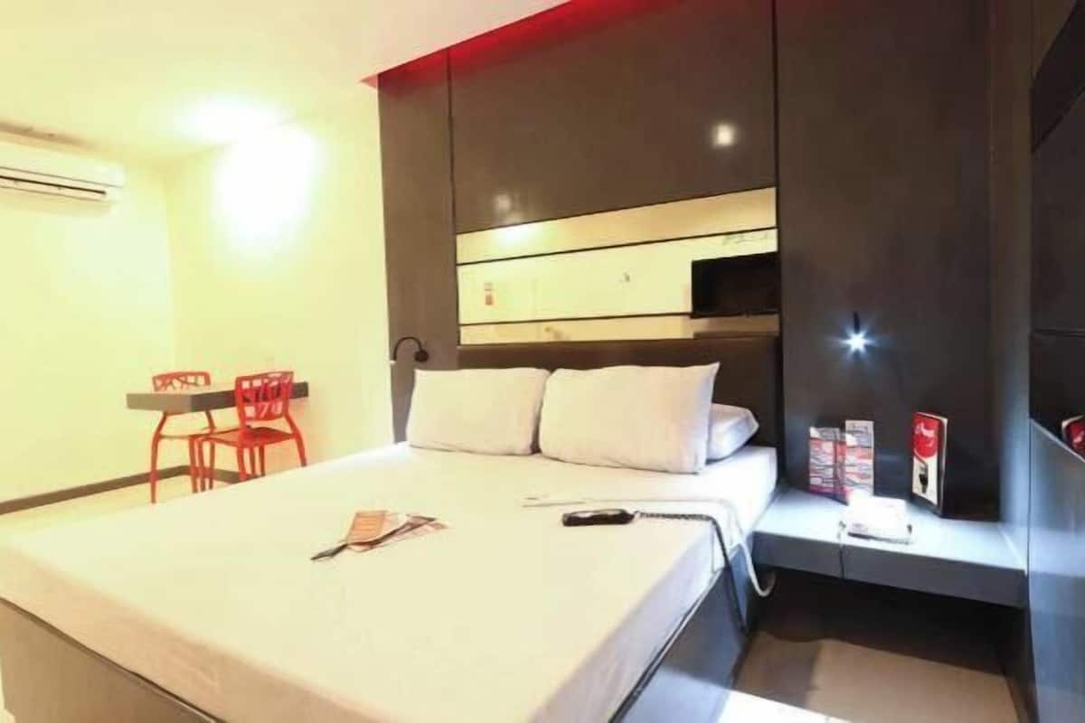 A hotel room with a bed, a desk, a chair, a TV, and a bedside table with a remote control.
