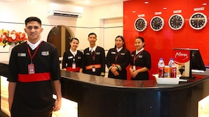 Ein Hotel-Empfangsbereich mit einer roten Wand, auf der Uhren für Manila, Tokio, Sydney und London angebracht sind. Vier Mitarbeiter stehen hinter dem Empfangstresen, der einen Menüständer mit Getränken hat.