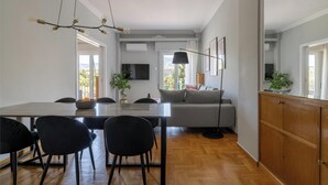 Ein modernes Wohnzimmer mit einer Couch, einem Esstisch und Stühlen. Ein Fernseher ist an der Wand befestigt, ein großes Fenster bietet einen Ausblick und eine Deckenleuchte sorgt für Licht.