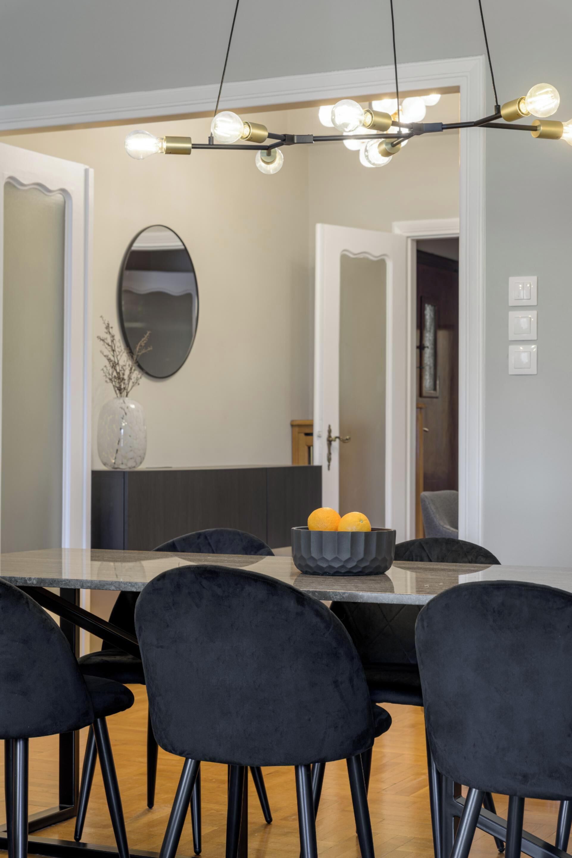A dining area with a modern chandelier, a round mirror, and a vase with branches.