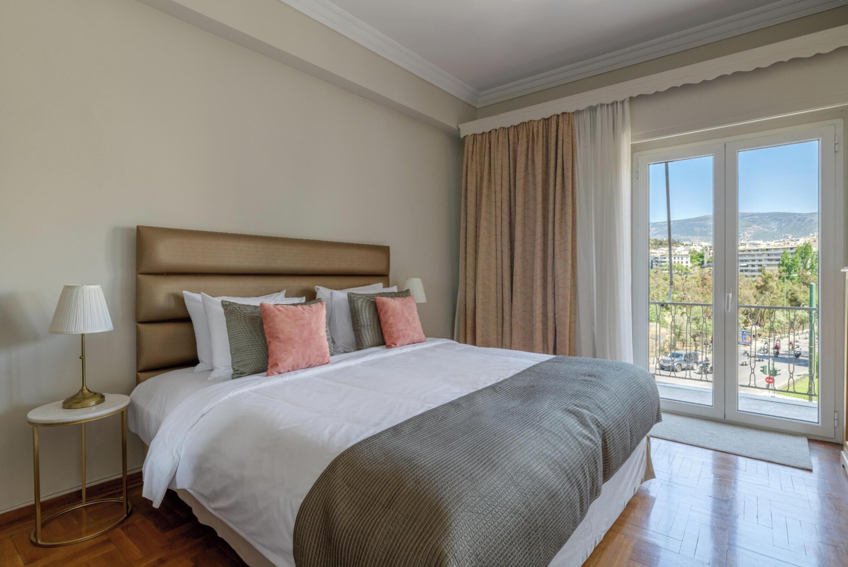 A hotel room with a large bed, a bedside lamp, and a view of the city through a large window.