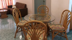 A dining area with a glass-top table surrounded by four wooden chairs with patterned cushions. A brown sofa and a small round table are visible in the background. - Glory by Alpine (Port Dickson)