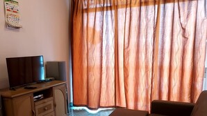A living room with a sofa, a television on a wooden cabinet, a wicker chair, and a window with orange curtains. - Glory by Alpine (Port Dickson)