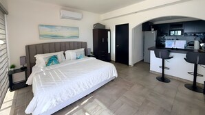 Une chambre moderne avec un grand lit, une petite table de chevet, une commode, un tableau, une armoire et un coin cuisine avec un bar.