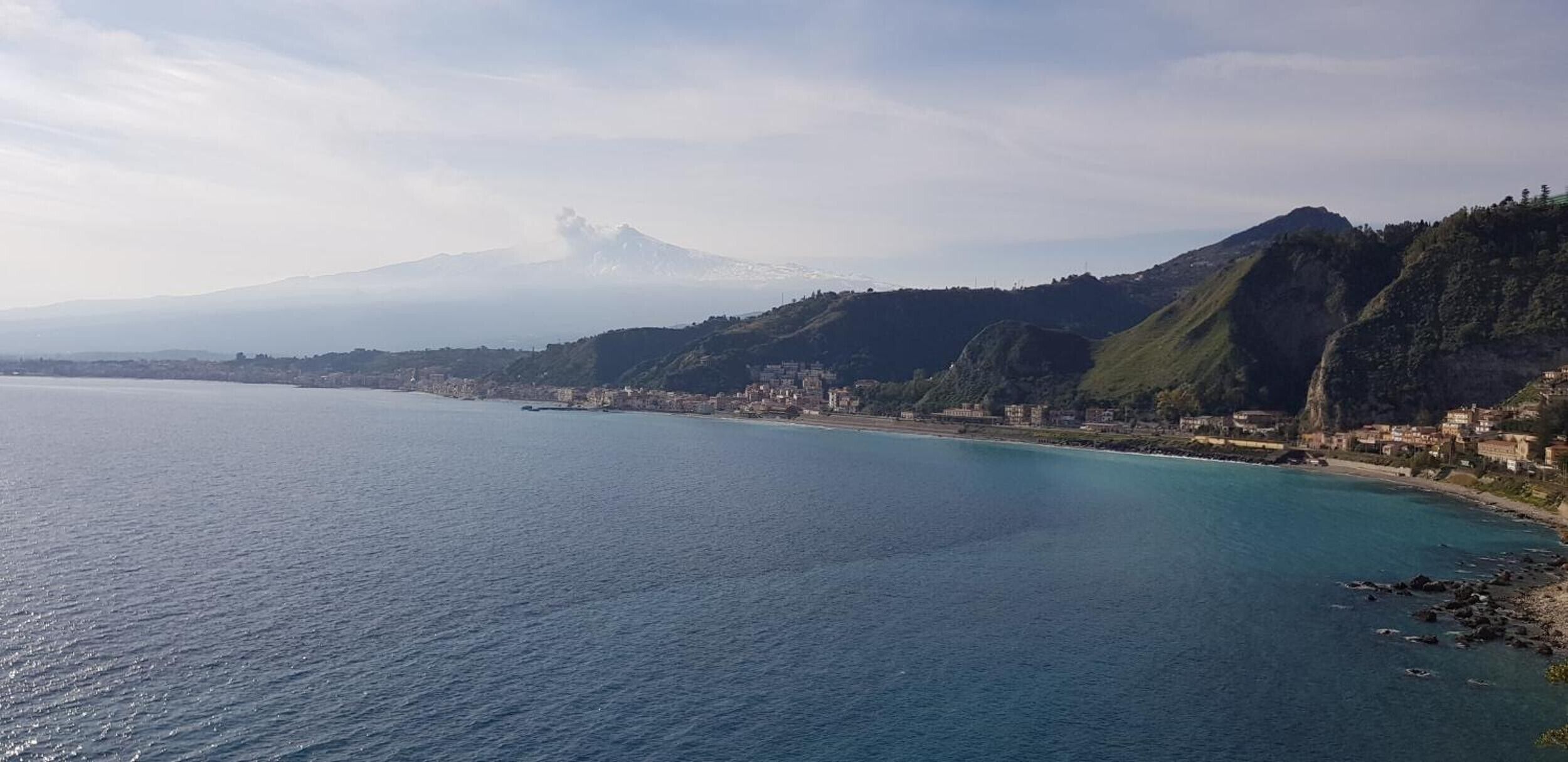 Eine Küstenstadt mit einem Berg im Hintergrund, ruhigem Meer und Gebäuden entlang der Uferlinie.