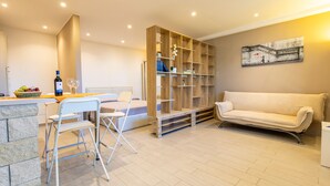 A modern living room with a sofa, a dining area with chairs, a wooden bookshelf, and a painting on the wall.