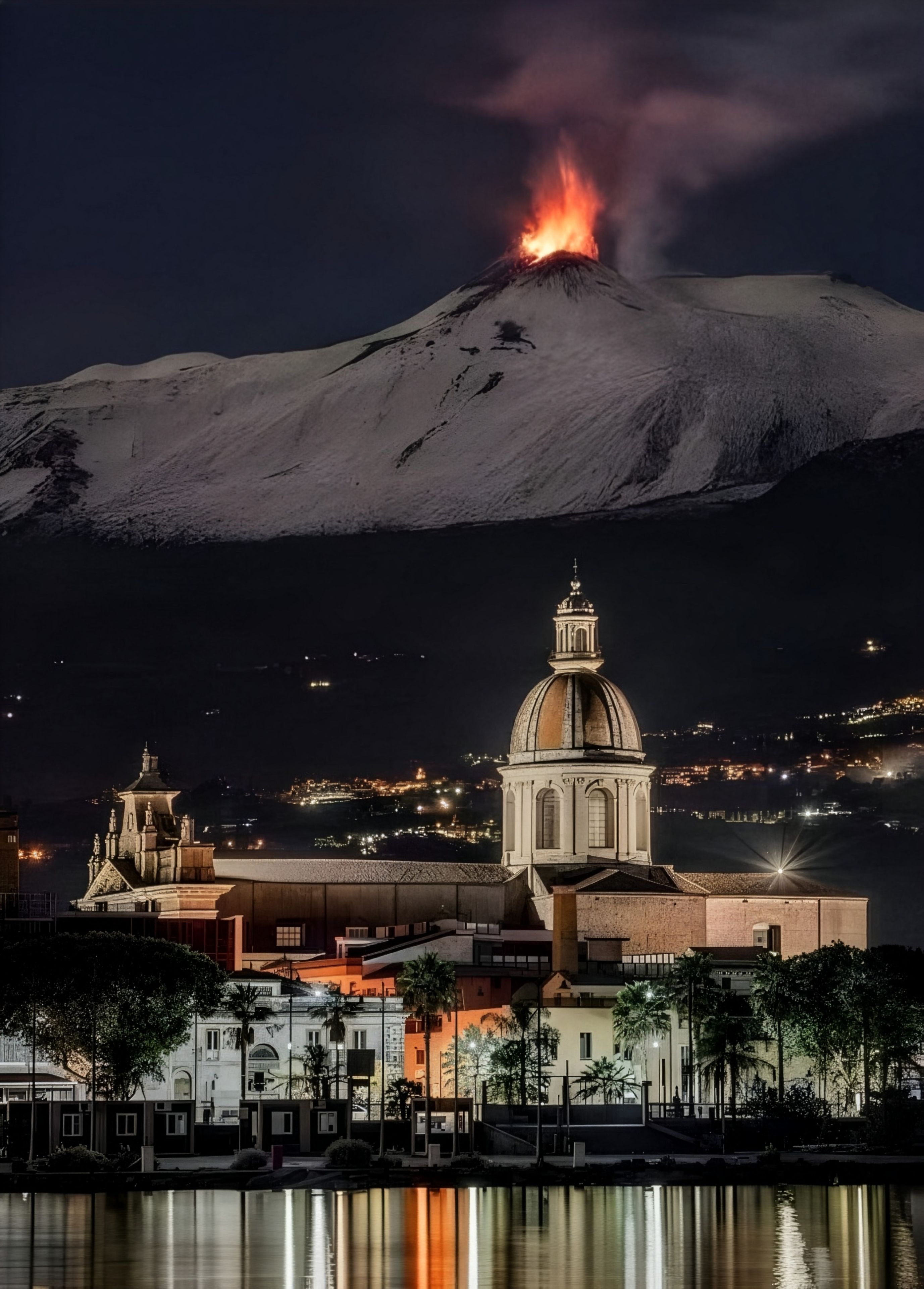 Una vista notturna di una città con un vulcano sullo sfondo, un edificio a cupola ben visibile e un'acqua calma che riflette le luci della città.