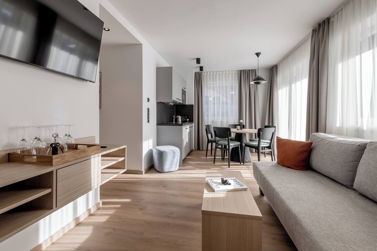 A modern living room with a flat-screen TV, a wooden coffee table, a grey sofa, and a dining area with a table and chairs.