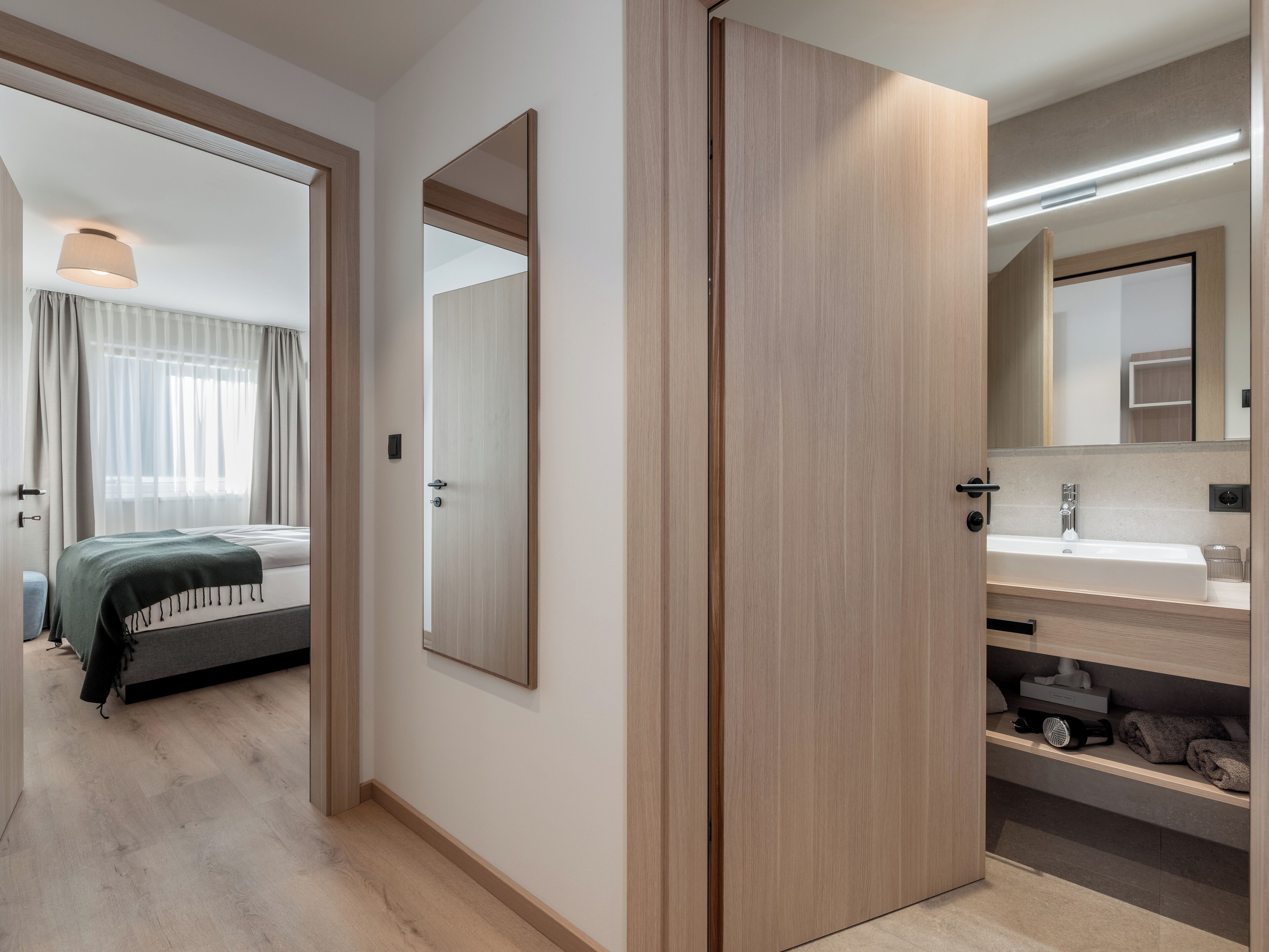 A modern hotel room with a bed, a bathroom with a sink and mirror, and a hallway leading to another room.