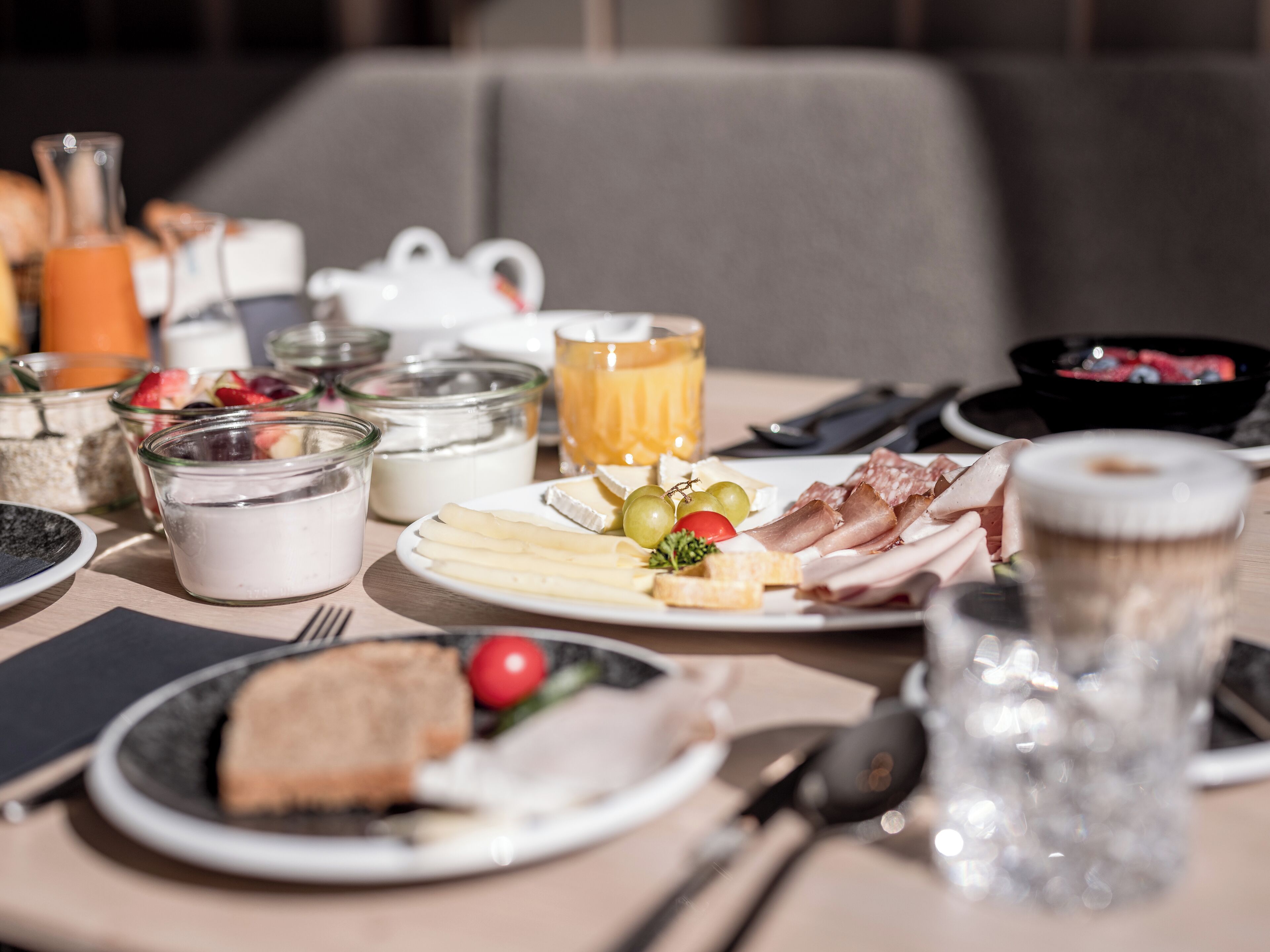 Ein Frühstückstisch mit verschiedenen Speisen, darunter Käse, Fleisch und Obst, mit einem Glas Saft und einer Teekanne.