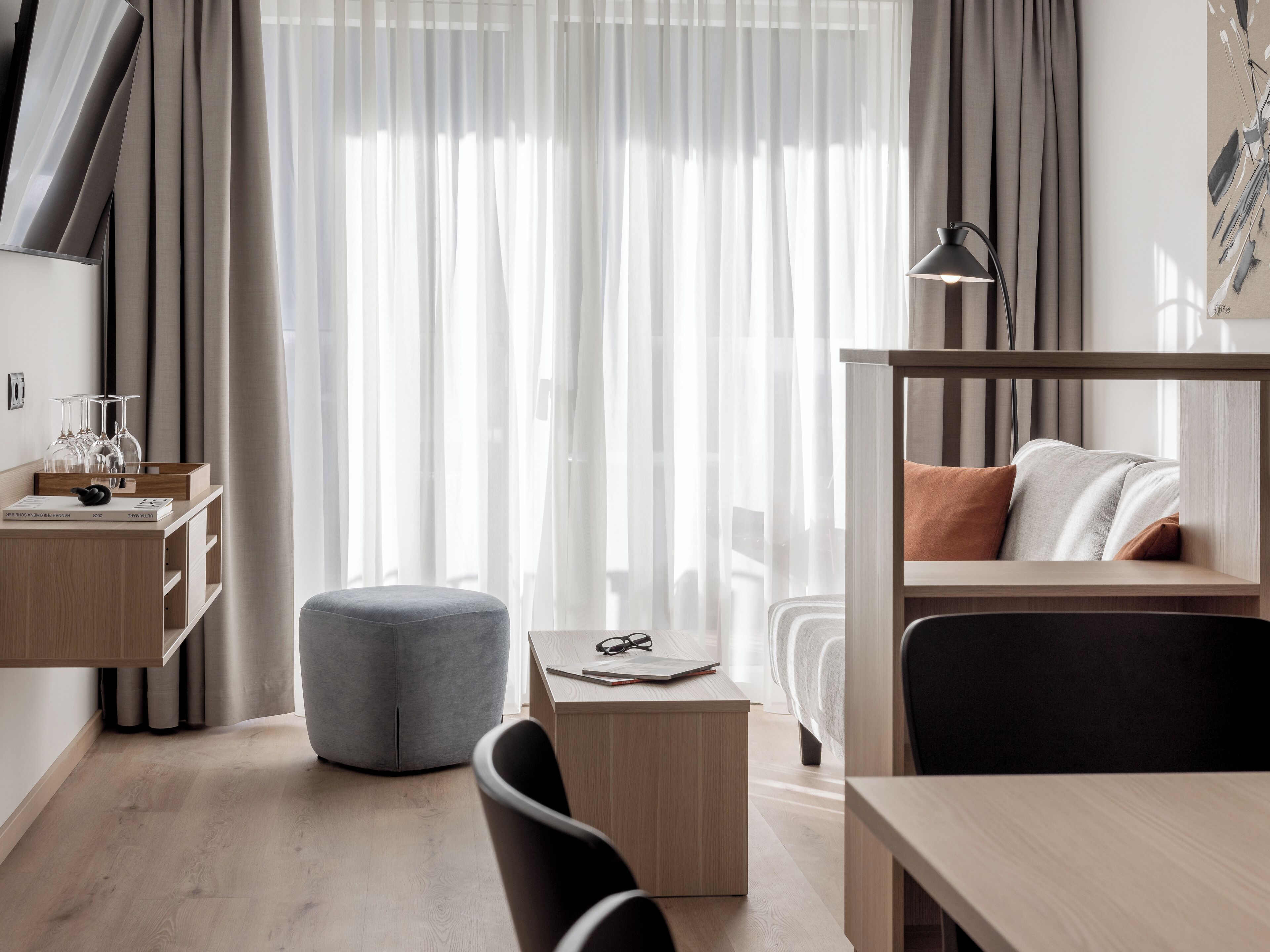 Une pièce moderne et bien éclairée, avec un bureau en bois, un fauteuil confortable, un petit pouf, un lit avec une tête de lit et une table de chevet avec une lampe.
