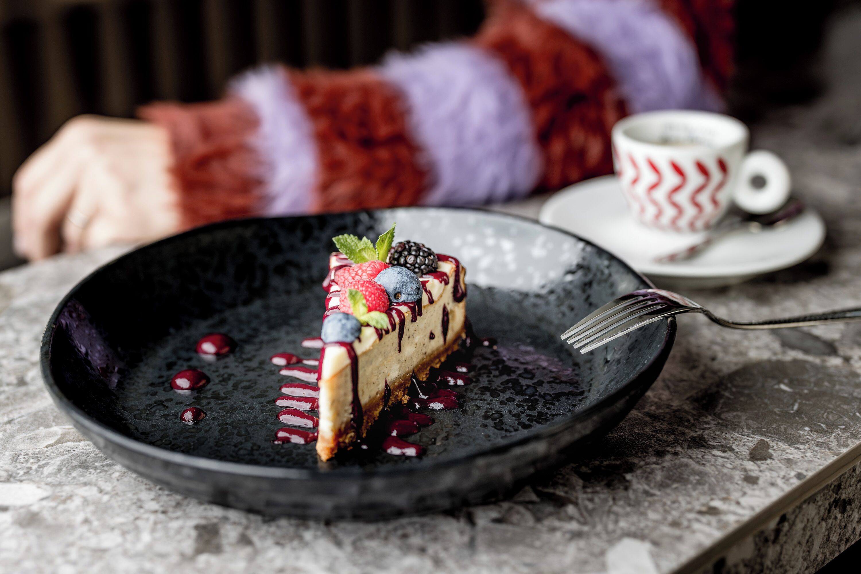 A slice of cheesecake with berries on a black plate, accompanied by a cup and saucer on a marble surface.
