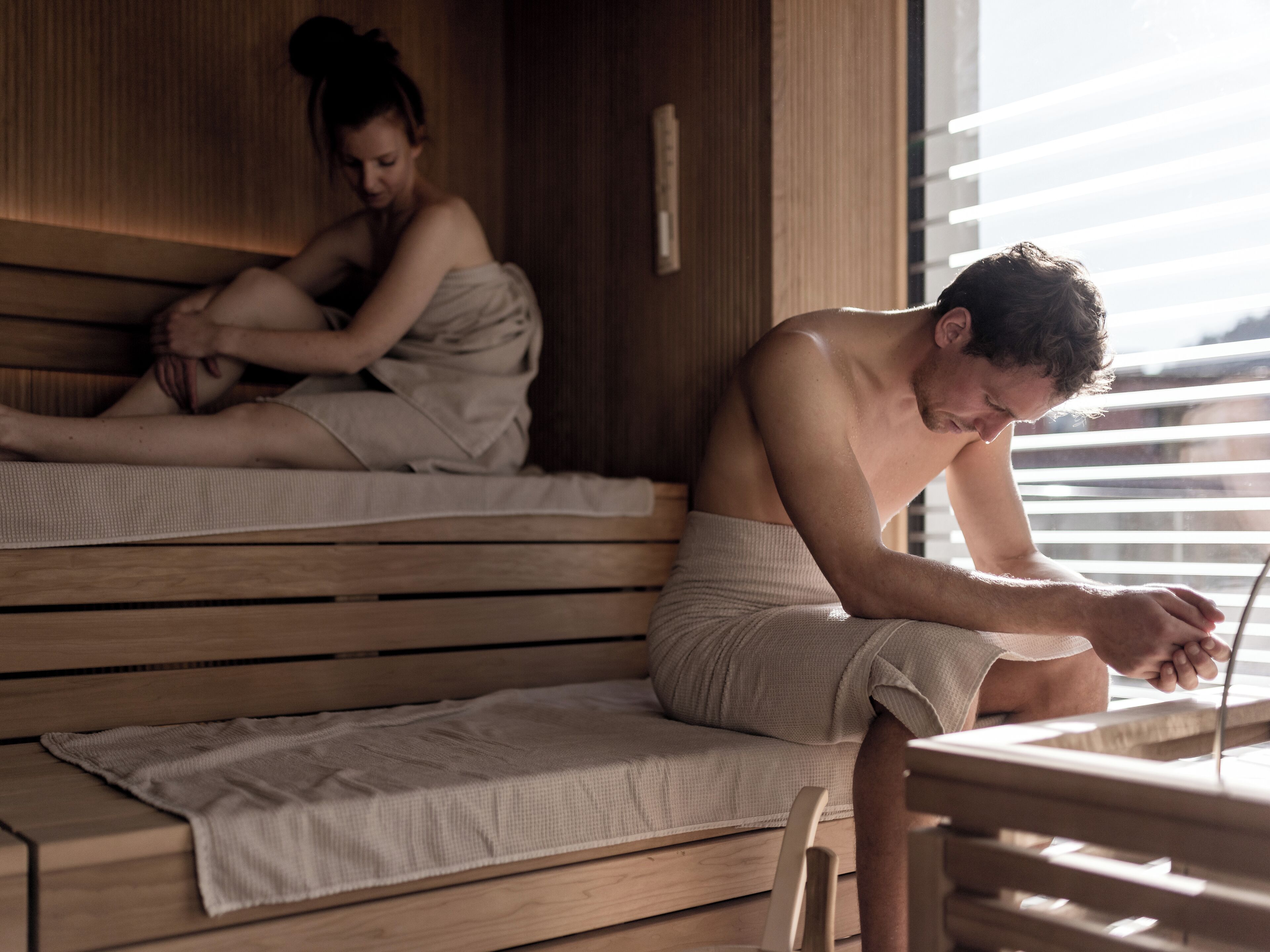 A wooden sauna with benches, a bench with a towel, and a window with blinds.